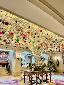 A festive indoor scene with a large, intricately decorated tree-like structure adorned with numerous colorful ornaments and sparkling lights. The room features ornate furniture, elegant chandeliers, and additional Christmas decorations such as garlands and a Christmas tree in the background.