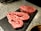 Two slices of marbled raw beef steak, possibly ribeye, are placed on a black slate surface. The beef is heavily marbled with fat, showcasing a mix of deep red meat and white fat. There are small flecks of seasoning, possibly pepper, on the surface of the meat. The background is a textured, speckled gray countertop with some kitchen tools blurred in the distance.