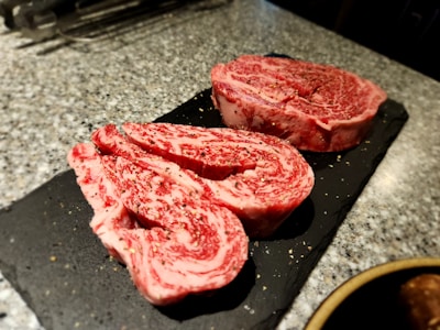 Two slices of marbled raw beef steak, possibly ribeye, are placed on a black slate surface. The beef is heavily marbled with fat, showcasing a mix of deep red meat and white fat. There are small flecks of seasoning, possibly pepper, on the surface of the meat. The background is a textured, speckled gray countertop with some kitchen tools blurred in the distance.