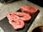 Two slices of marbled raw beef steak, possibly ribeye, are placed on a black slate surface. The beef is heavily marbled with fat, showcasing a mix of deep red meat and white fat. There are small flecks of seasoning, possibly pepper, on the surface of the meat. The background is a textured, speckled gray countertop with some kitchen tools blurred in the distance.