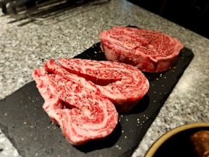 Two slices of marbled raw beef steak, possibly ribeye, are placed on a black slate surface. The beef is heavily marbled with fat, showcasing a mix of deep red meat and white fat. There are small flecks of seasoning, possibly pepper, on the surface of the meat. The background is a textured, speckled gray countertop with some kitchen tools blurred in the distance.