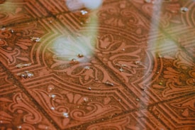 A wet decorative tile floor with an intricate floral and geometric pattern. The surface is covered with small puddles and droplets of water, reflecting some light.