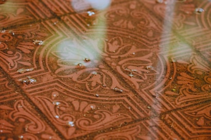 A wet decorative tile floor with an intricate floral and geometric pattern. The surface is covered with small puddles and droplets of water, reflecting some light.