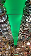 A room filled with bicycles and bicycle parts, with rows of bicycle wheels hanging from the ceiling. The ceiling is illuminated with a green light, creating a dramatic effect throughout the room. The space is densely packed with various bike components, reflecting a cluttered yet organized environment typical of a bike shop or a repair area.