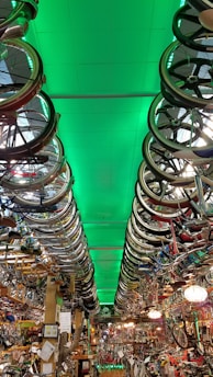 A room filled with bicycles and bicycle parts, with rows of bicycle wheels hanging from the ceiling. The ceiling is illuminated with a green light, creating a dramatic effect throughout the room. The space is densely packed with various bike components, reflecting a cluttered yet organized environment typical of a bike shop or a repair area.