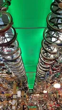 A room filled with bicycles and bicycle parts, with rows of bicycle wheels hanging from the ceiling. The ceiling is illuminated with a green light, creating a dramatic effect throughout the room. The space is densely packed with various bike components, reflecting a cluttered yet organized environment typical of a bike shop or a repair area.