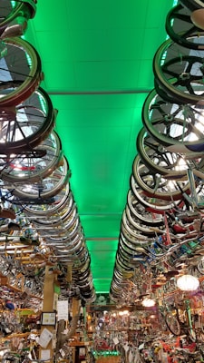 A room filled with bicycles and bicycle parts, with rows of bicycle wheels hanging from the ceiling. The ceiling is illuminated with a green light, creating a dramatic effect throughout the room. The space is densely packed with various bike components, reflecting a cluttered yet organized environment typical of a bike shop or a repair area.