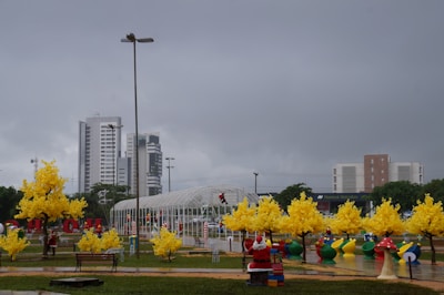 A cityscape featuring tall modern buildings under a cloudy sky. Several ornamental yellow trees and colorful playground equipment are scattered across a wet, glossy surface in a park setting. There are festive decorations, including images of Santa Claus and candy canes.