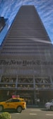 A tall skyscraper with a modern glass and steel facade extends upwards against a backdrop of a blue sky with scattered thin clouds. At the base of the building, the name 'The New York Times' is prominently displayed. A yellow taxi and a white vehicle are visible on the street in the foreground.