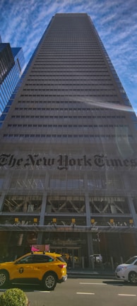 A tall skyscraper with a modern glass and steel facade extends upwards against a backdrop of a blue sky with scattered thin clouds. At the base of the building, the name 'The New York Times' is prominently displayed. A yellow taxi and a white vehicle are visible on the street in the foreground.
