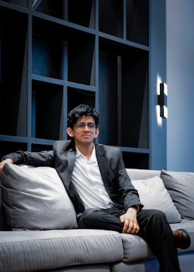 A man in a black suit and white shirt is seated on a light gray sofa with a contemporary design. The background features a modern black shelving unit with geometric patterns and a sleek wall lamp on the side. The setting appears to be an upscale, minimalist living space with a calm and professional ambiance.