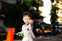 A young child is pointing towards the camera while holding a small green toy in an outdoor setting. The child is wearing a light-colored shirt with cartoon motifs. In the background, there are benches, trees, and colorful child-sized ride-on toys.