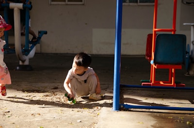 A young child wearing a miniature hardhat and denim overalls, playing with a toy dump truck in a backyard.