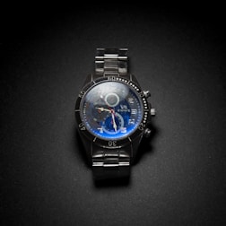 A metallic wristwatch with a dark face featuring large numerals and three small dials for additional functions. The watch has a sturdy metal bracelet, and the face has a blue tint.