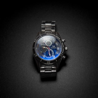 A metallic wristwatch with a dark face featuring large numerals and three small dials for additional functions. The watch has a sturdy metal bracelet, and the face has a blue tint.