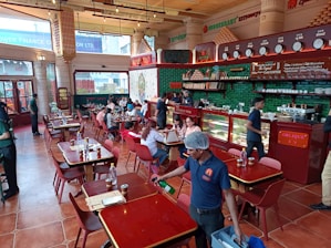 A bustling restaurant interior with people dining at wooden tables. The space is adorned with red and green accents, featuring a counter displaying various items with staff attending to customers. The atmosphere is lively with patrons engaged in conversations, and a server cleaning near the front.