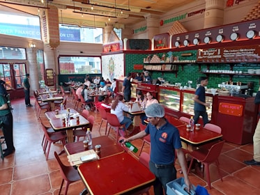 A bustling restaurant interior with people dining at wooden tables. The space is adorned with red and green accents, featuring a counter displaying various items with staff attending to customers. The atmosphere is lively with patrons engaged in conversations, and a server cleaning near the front.