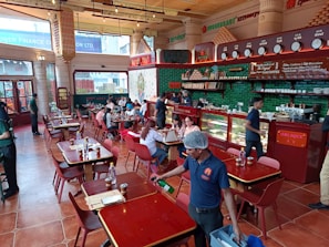 A bustling restaurant interior with people dining at wooden tables. The space is adorned with red and green accents, featuring a counter displaying various items with staff attending to customers. The atmosphere is lively with patrons engaged in conversations, and a server cleaning near the front.