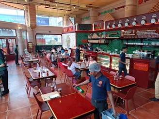 A bustling restaurant interior with people dining at wooden tables. The space is adorned with red and green accents, featuring a counter displaying various items with staff attending to customers. The atmosphere is lively with patrons engaged in conversations, and a server cleaning near the front.