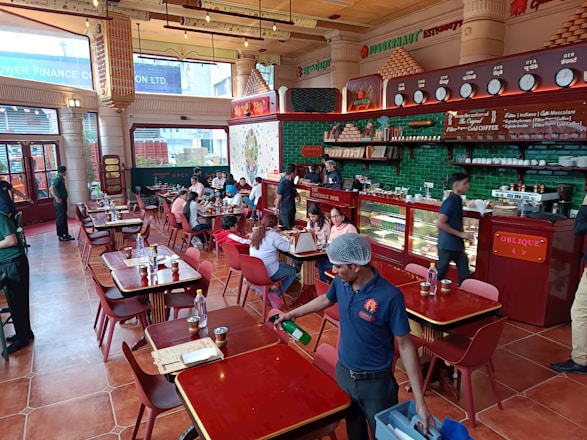 A bustling restaurant interior with people dining at wooden tables. The space is adorned with red and green accents, featuring a counter displaying various items with staff attending to customers. The atmosphere is lively with patrons engaged in conversations, and a server cleaning near the front.