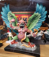 A colorful statue depicting a mythical creature with large, detailed wings in shades of blue and green. The creature has a pink humanoid torso with horns and an animal-like face that resembles a bird's beak. It holds snakes in its hands and has more snakes coiled around its body. Its legs are green and resemble those of a bird, ending in red talons. The statue is placed on a black base.