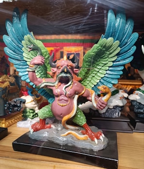 A colorful statue depicting a mythical creature with large, detailed wings in shades of blue and green. The creature has a pink humanoid torso with horns and an animal-like face that resembles a bird's beak. It holds snakes in its hands and has more snakes coiled around its body. Its legs are green and resemble those of a bird, ending in red talons. The statue is placed on a black base.