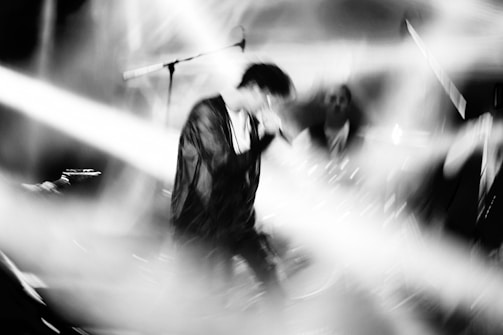A blurred black and white image capturing the movement and energy of a musician performing on stage. Bright stage lights create dynamic lines across the scene, adding to the sense of motion.