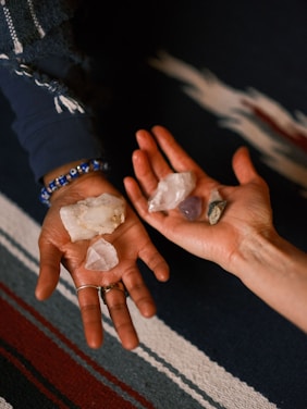 Two hands are holding a collection of crystals of various shapes and colors, positioned above a textured, multicolored fabric. The left hand, adorned with a beaded bracelet and rings, holds larger, white crystals, while the right hand holds smaller, varied stones.