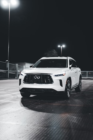 A white SUV is parked under bright lights in a dimly lit area. The vehicle has a sleek design with a prominent front grille and modern headlamps. The setting appears to be a parking lot at night, with additional lights illuminating the background.