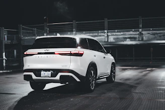 A white Infiniti QX60 is parked in a dimly lit, empty parking area. The vehicle is positioned at an angle, showing its rear and side. The environment is industrial, with metal railings and concrete structures in the background. The lighting creates reflections on the car's surface and the wet ground.