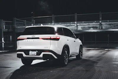 A white Infiniti QX60 is parked in a dimly lit, empty parking area. The vehicle is positioned at an angle, showing its rear and side. The environment is industrial, with metal railings and concrete structures in the background. The lighting creates reflections on the car's surface and the wet ground.
