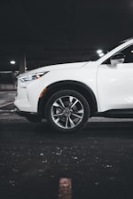 A white SUV is parked in a dimly lit underground parking garage. The focus is on the front wheel and the side of the vehicle, highlighting its sleek design and modern alloy wheels. The setting has an industrial feel with concrete flooring and muted lighting.