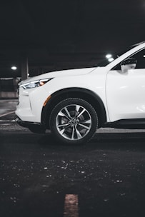 A white SUV is parked in a dimly lit underground parking garage. The focus is on the front wheel and the side of the vehicle, highlighting its sleek design and modern alloy wheels. The setting has an industrial feel with concrete flooring and muted lighting.