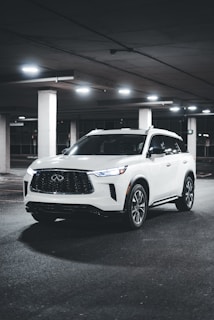 A white SUV is parked in an underground parking lot with dim lighting. The vehicle's headlights are off, and the surroundings are concrete with several pillars and overhead lights.