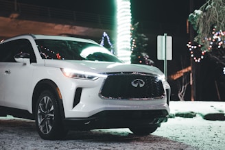 A sleek, white luxury SUV is parked in a snowy outdoor setting at night. The vehicle is prominently lit, reflecting the glow of surrounding lights. Bright, festive decorations and strings of lights are visible in the background, creating a colorful and lively ambiance. Snow is lightly covering the ground around the car.