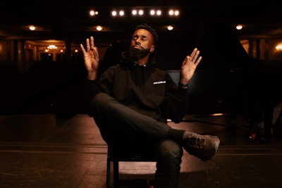 A person sits cross-legged on a chair, with eyes closed, hands raised, and a calm expression in a dimly lit auditorium. The surroundings include a stage, overhead lights, and a grand piano partially visible in the background.
