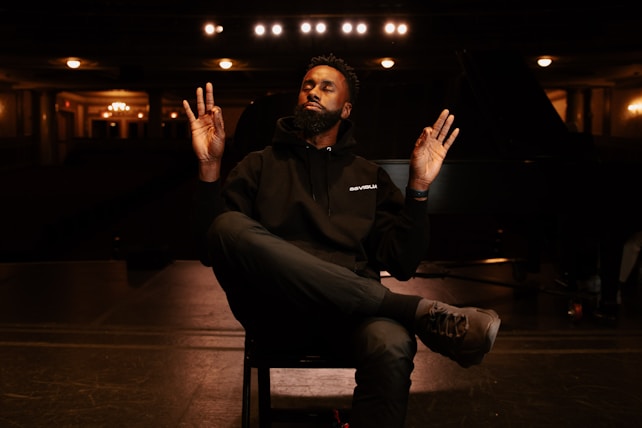 A person sits cross-legged on a chair, with eyes closed, hands raised, and a calm expression in a dimly lit auditorium. The surroundings include a stage, overhead lights, and a grand piano partially visible in the background.