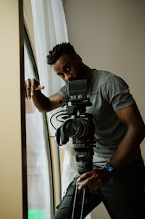A person stands beside a professional video camera mounted on a tripod. The individual is casually dressed in a gray t-shirt and appears to be indoors near a window with white curtains.