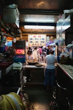 A bustling market stall filled with various items and people. There's a prominent sign with Chinese characters, and people are gathered around, engaging in different activities. Cooking equipment and various hanging items are visible, contributing to a busy and lively atmosphere.