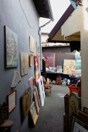 An alleyway lined with various framed paintings hanging and leaning against a wall, creating an art gallery atmosphere. A wooden chair is placed in the center, and several other objects like vases and decorative items are arranged on a table at the far end.