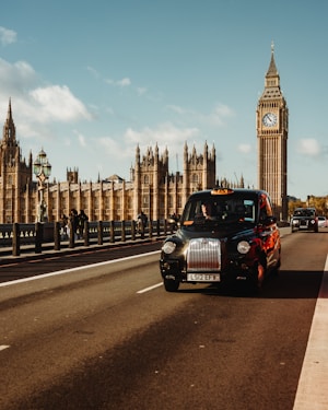 A classic black taxi drives on a bridge with the Palace of Westminster and Big Ben in the background. The sky is clear with a few clouds. The architectural details and the iconic clock tower are prominent. Several people can be seen walking along the bridge.
