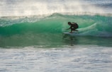 A surfer skillfully navigates a translucent green wave, crouched low on his board as spray shimmers in the sunlight. The waves roll in the background, creating a dynamic scene full of movement and energy.