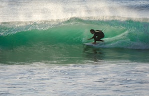A surfer skillfully navigates a translucent green wave, crouched low on his board as spray shimmers in the sunlight. The waves roll in the background, creating a dynamic scene full of movement and energy.