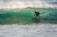 A surfer skillfully navigates a translucent green wave, crouched low on his board as spray shimmers in the sunlight. The waves roll in the background, creating a dynamic scene full of movement and energy.