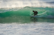 A surfer skillfully navigates a translucent green wave, crouched low on his board as spray shimmers in the sunlight. The waves roll in the background, creating a dynamic scene full of movement and energy.