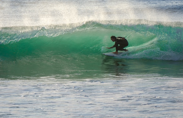 A surfer skillfully navigates a translucent green wave, crouched low on his board as spray shimmers in the sunlight. The waves roll in the background, creating a dynamic scene full of movement and energy.
