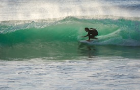 A surfer skillfully navigates a translucent green wave, crouched low on his board as spray shimmers in the sunlight. The waves roll in the background, creating a dynamic scene full of movement and energy.