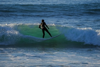 A surfer in a wetsuit rides a wave, maintaining balance while navigating through the surf. The wave is curling around with a striking green hue, and the ocean appears expansive under the natural light.