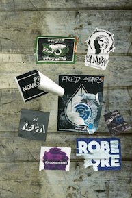 Various stickers are placed on a wooden surface, including designs with text such as 'Fixed Stars,' 'Bolognastickers,' and 'Tattoo Studio.' Some stickers show wear and tear, with edges peeling or faded areas. The background consists of rustic wooden planks with visible grain and weathering.