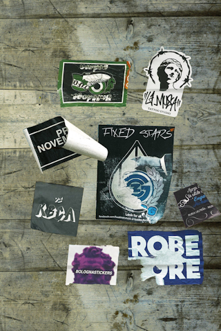 Various stickers are placed on a wooden surface, including designs with text such as 'Fixed Stars,' 'Bolognastickers,' and 'Tattoo Studio.' Some stickers show wear and tear, with edges peeling or faded areas. The background consists of rustic wooden planks with visible grain and weathering.